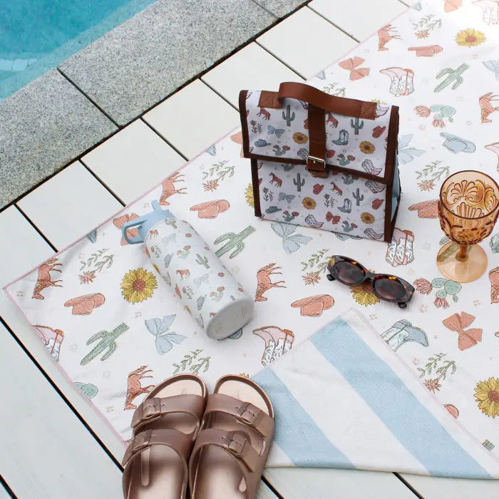 A styled flat-lay of a desert-themed canvas tote bag and matching accessories on a white tiled poolside deck. The tote bag has brown leather handles and trim, and the matching accessories include a cylindrical bag, a striped towel, sunglasses, and a glass, all featuring a cactus and sun motif.