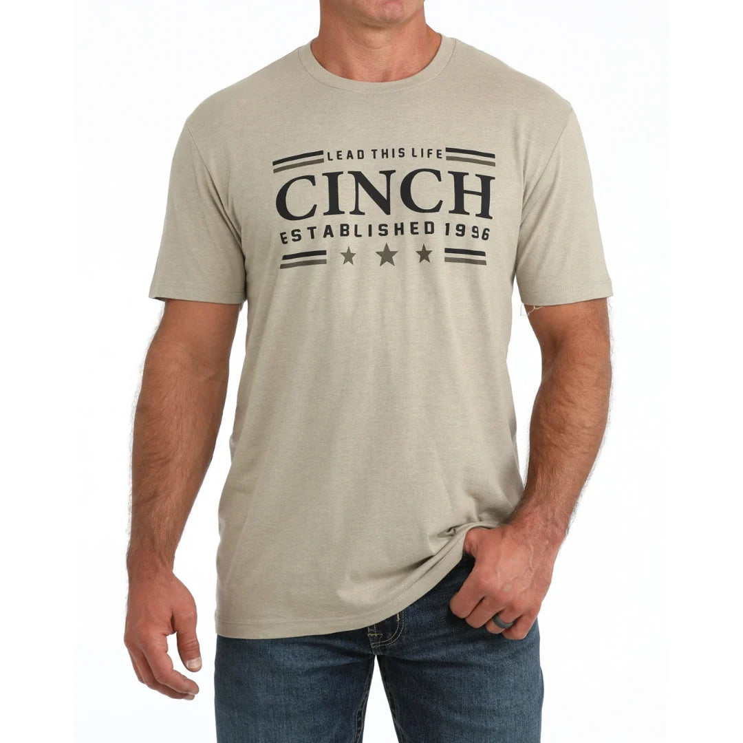 A man wearing a light beige short-sleeve t-shirt with a black graphic print on the front that reads 'LEAD THIS LIFE CINCH ESTABLISHED 1956'.