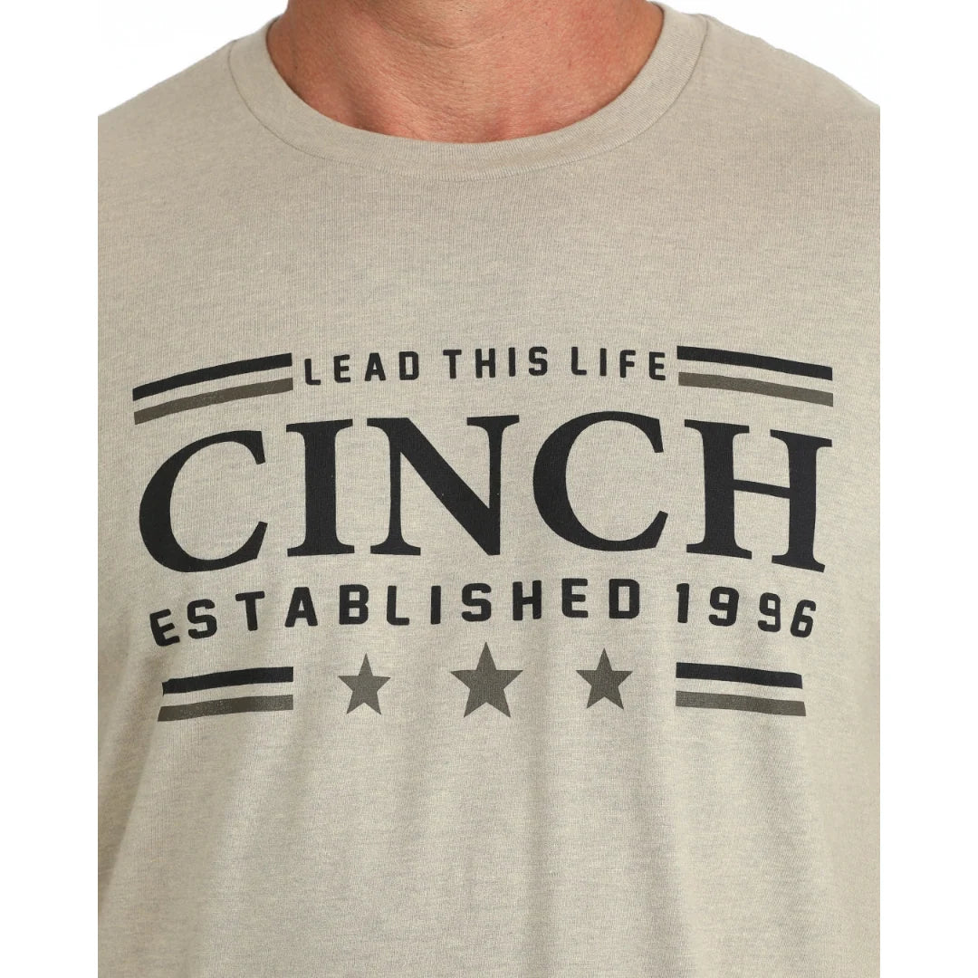 A close-up of a person wearing a light beige heather t-shirt with a dark gray graphic print that reads 'LEAD THIS LIFE CINCH ESTABLISHED 1996' with stars and stripes.