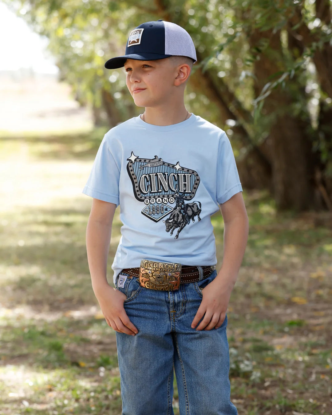 A young boy wearing a light blue graphic t-shirt with 'CINCH 2025' and a horse design, paired with blue jeans, a western belt, and a navy and white trucker hat, standing outdoors in a wooded area.