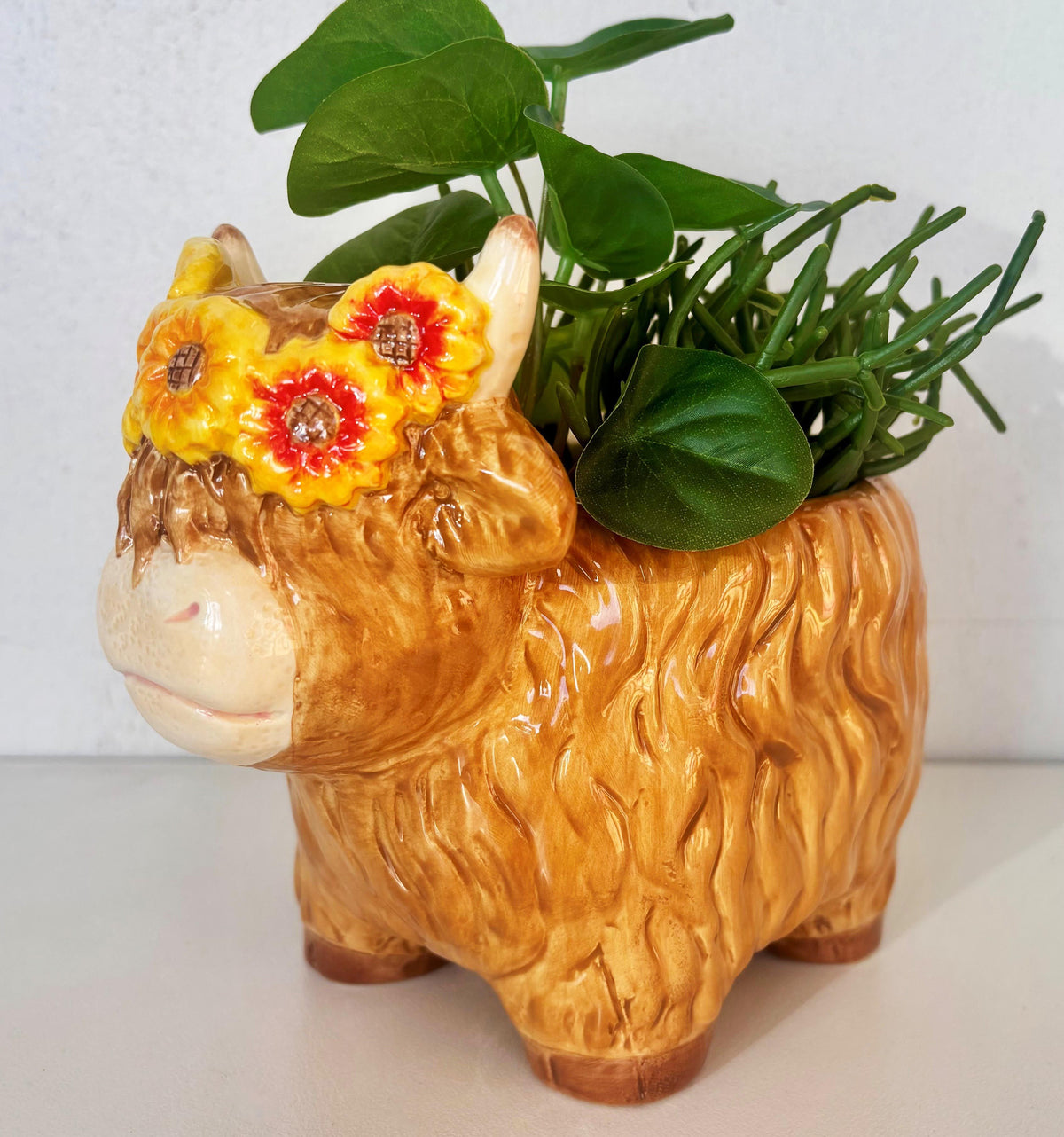 A ceramic highland cow planter in a warm golden-brown color, decorated with yellow and red flowers on its forehead and filled with green leafy plants, set against a plain light background.