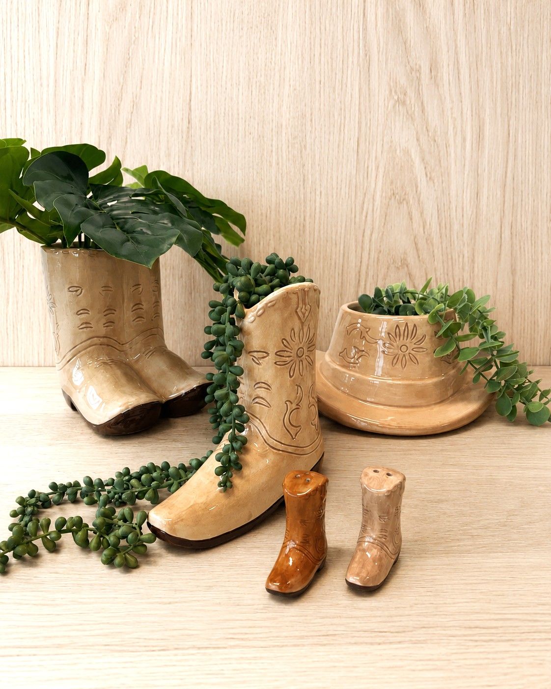 Three ceramic cowboy boot planters in beige and brown tones, decorated with engraved floral details and filled with green trailing plants, arranged on a light wood surface against a wood-paneled background.