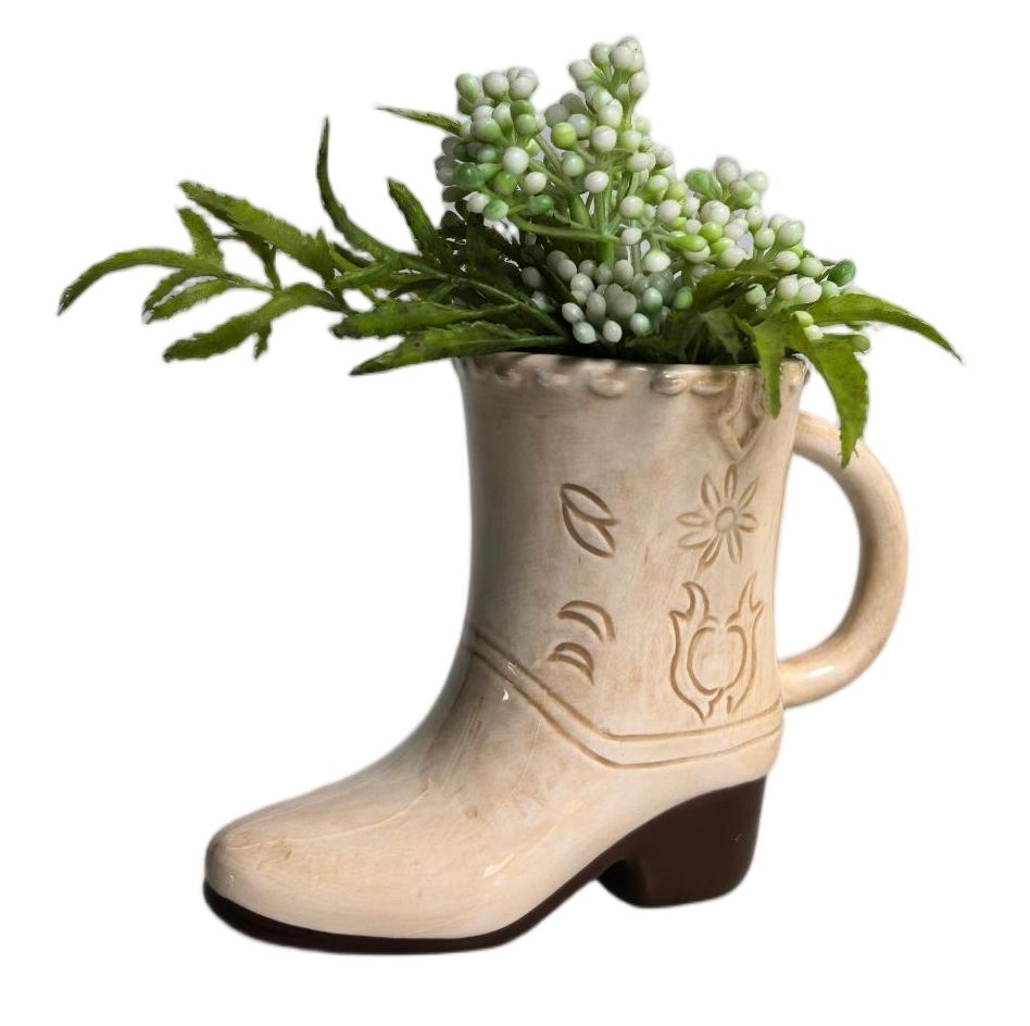 A ceramic cowboy boot planter in a cream color with a dark brown heel, filled with artificial green foliage and white berries, isolated on a white background.