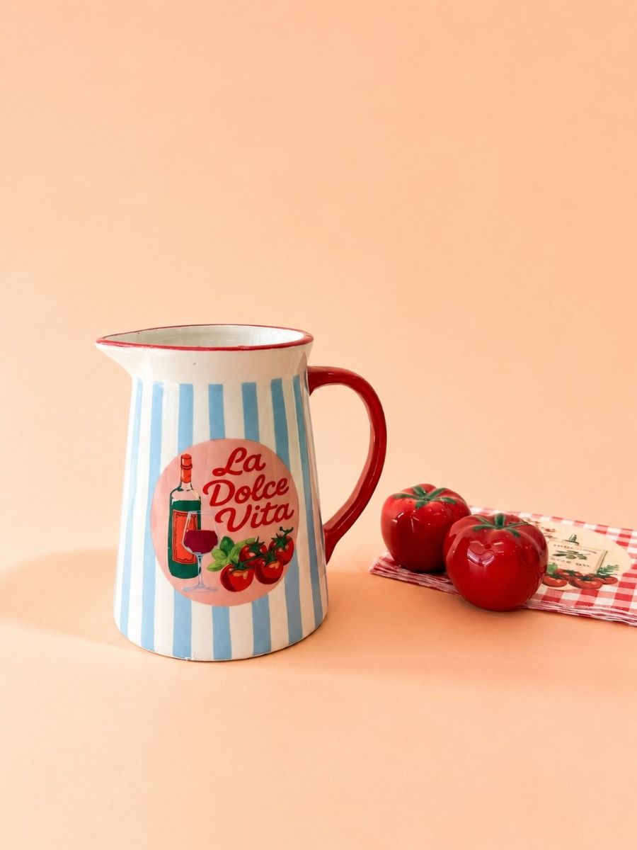 A ceramic pitcher with light blue and white vertical stripes, a red handle, and a red rim, featuring a retro circular 'La Dolce Vita' graphic. It is styled on a peach background next to two red tomatoes and a checkered napkin.