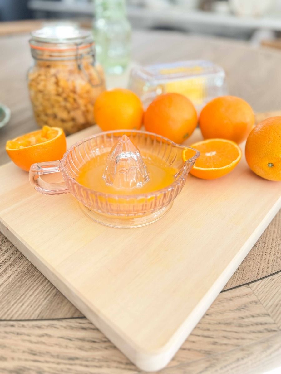 A clear glass citrus juicer with a handle sits on a light wooden cutting board, surrounded by fresh oranges and orange halves, with a jar of dried fruit in the softly blurred background.