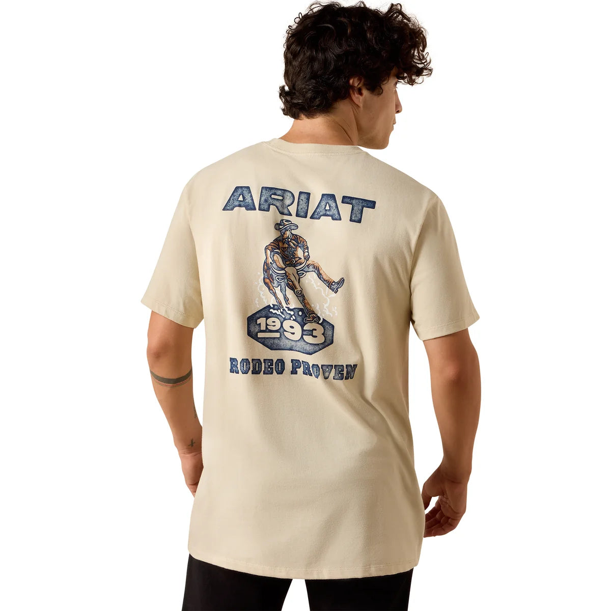 A man shown from the back wearing a cream-colored t-shirt with a large blue and brown graphic on the back that reads 'ARIAT 1993 RODEO PROVEN'.