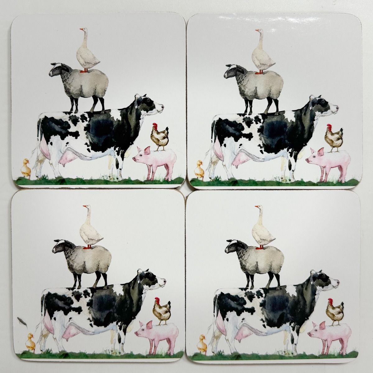 Four square coasters arranged in a 2x2 grid, each printed with a whimsical farm scene featuring a black-and-white cow, sheep, goose, piglets, chickens, and ducklings on a white background.