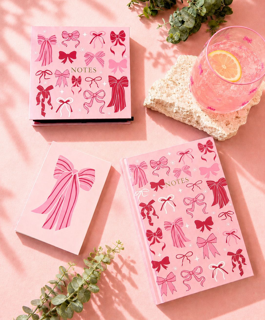 A pink notebook or journal featuring a stylized pink ribbon bow illustration on the cover, shown against a clean white background with a soft drop shadow.