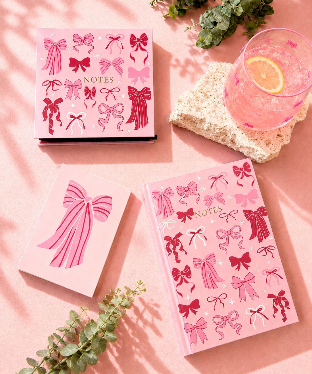 Pink notebook covers with bow illustrations arranged on a pink surface, styled with a glass of pink drink, a stone coaster, and greenery.