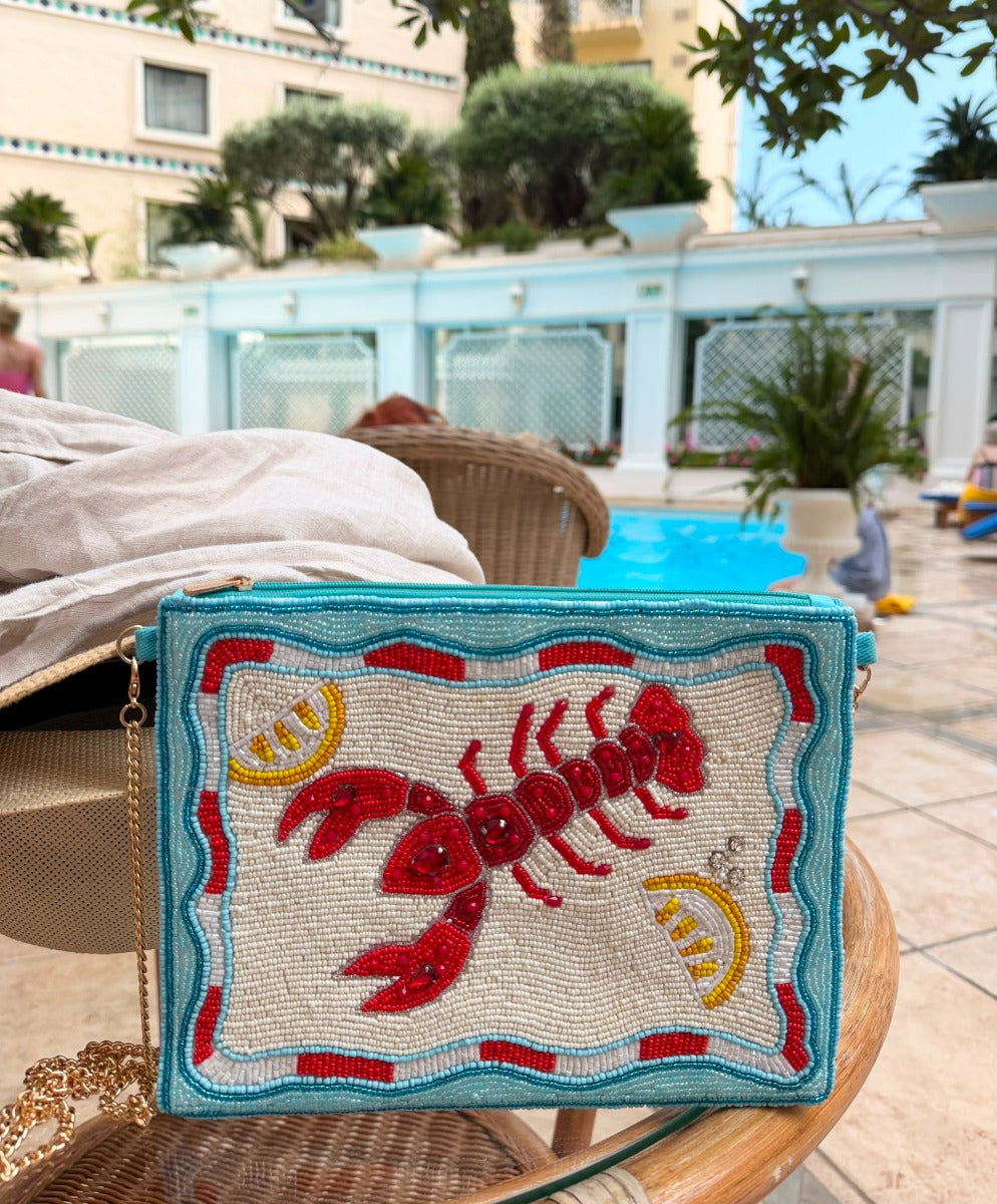 A rectangular clutch bag with a turquoise blue border and a raised red lobster design, decorated with yellow lemon slices and blue shell-like accents, resting on a wicker chair in an outdoor poolside setting.