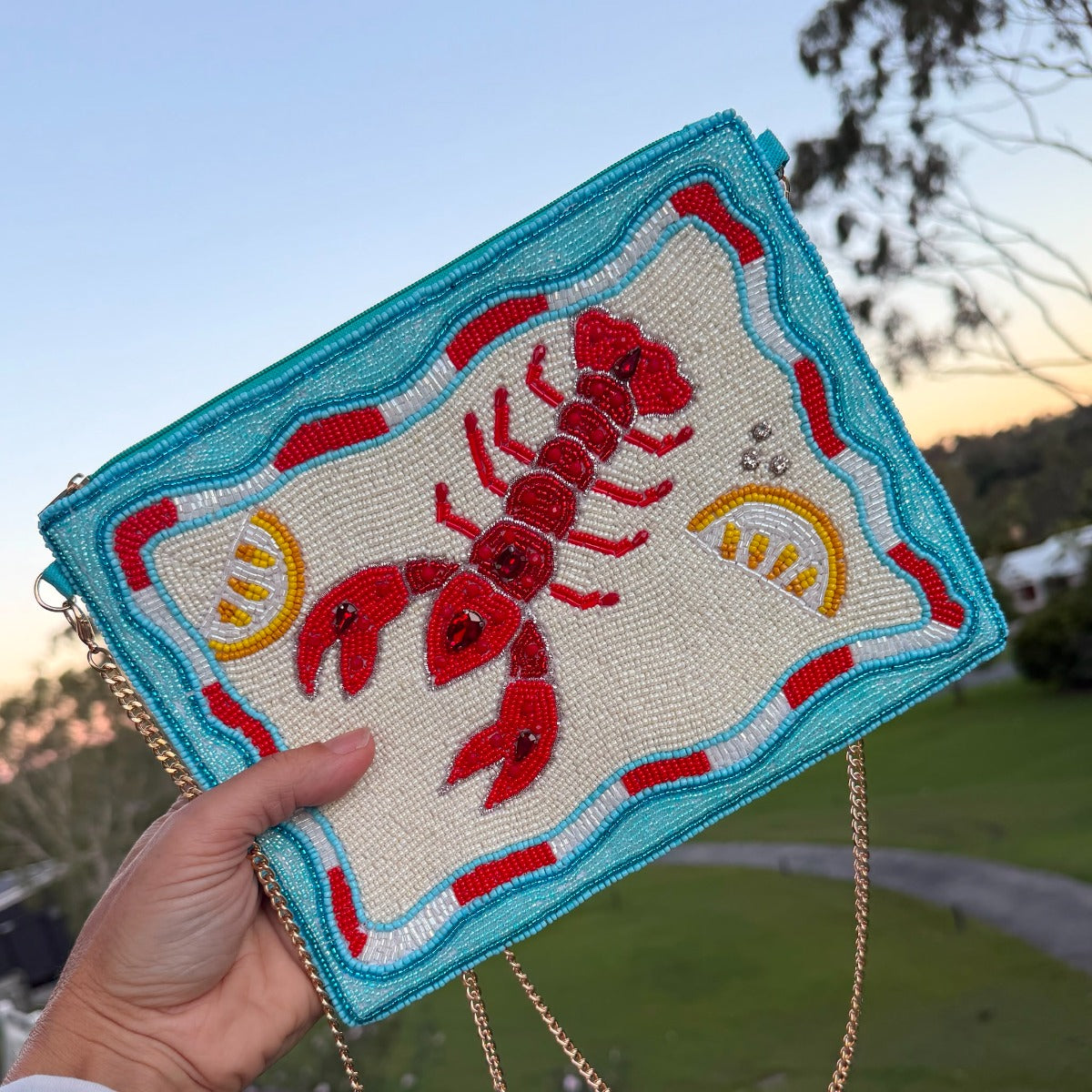 A rectangular clutch bag with a turquoise blue border and a raised red lobster design, decorated with yellow lemon slices and blue shell-like accents, resting on a wicker chair in an outdoor poolside setting.